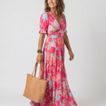 Model wearing a pink floral midi dress with short sleeves, V-neck, and tiered skirt, holding a tan tote bag.
