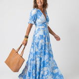 Blue V-neck midi dress with short puff sleeves, white floral print, and tiered skirt, worn with a brown tote.